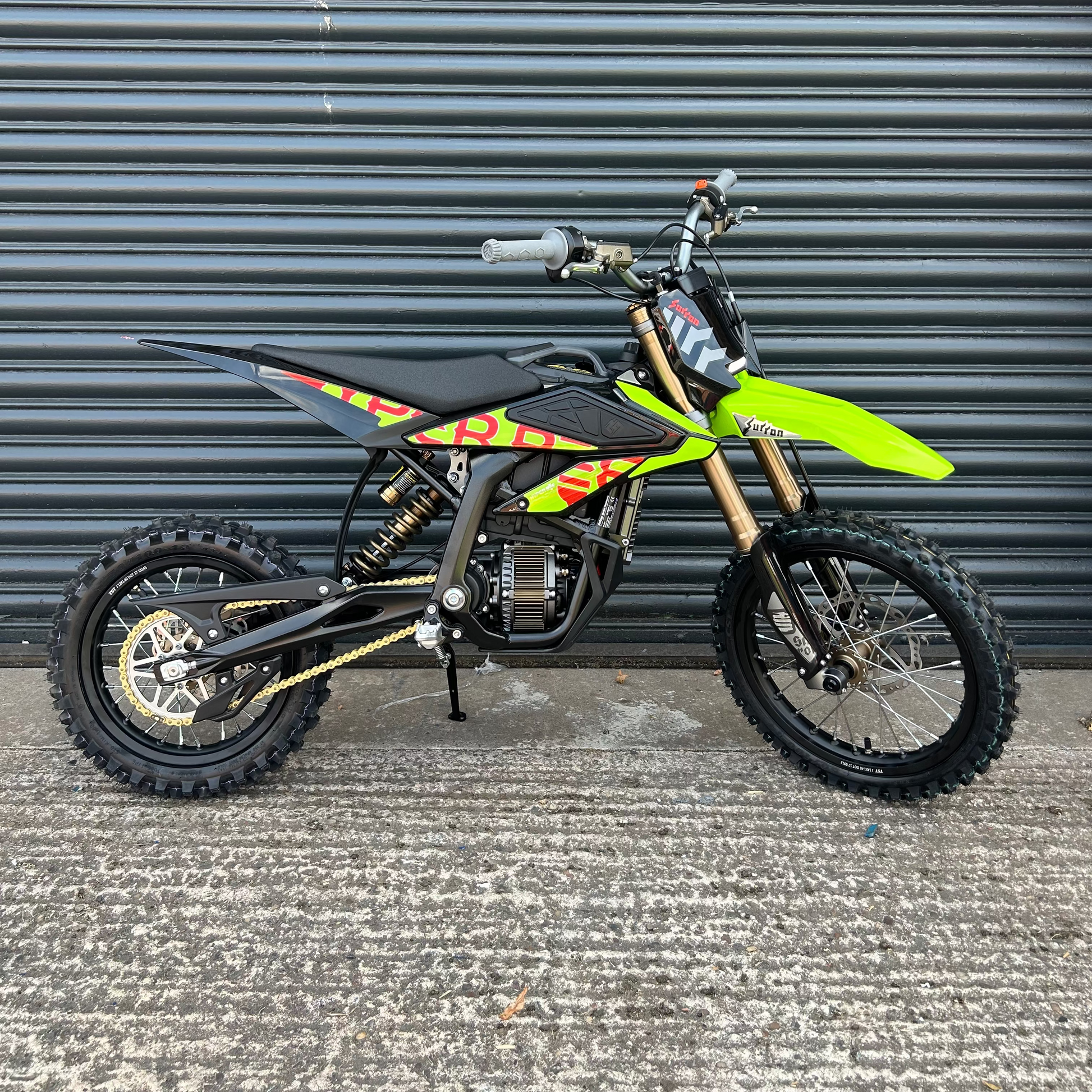 A small off-road dirt bike with a bright neon green front fender, black seat and body panels, and yellow/orange graphic decals, photographed against a dark gray metal roll-up door.
