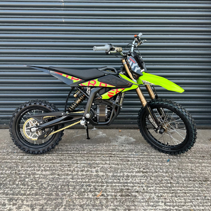A small off-road dirt bike with a bright neon green front fender, black seat and body panels, and yellow/orange graphic decals, photographed against a dark gray metal roll-up door.