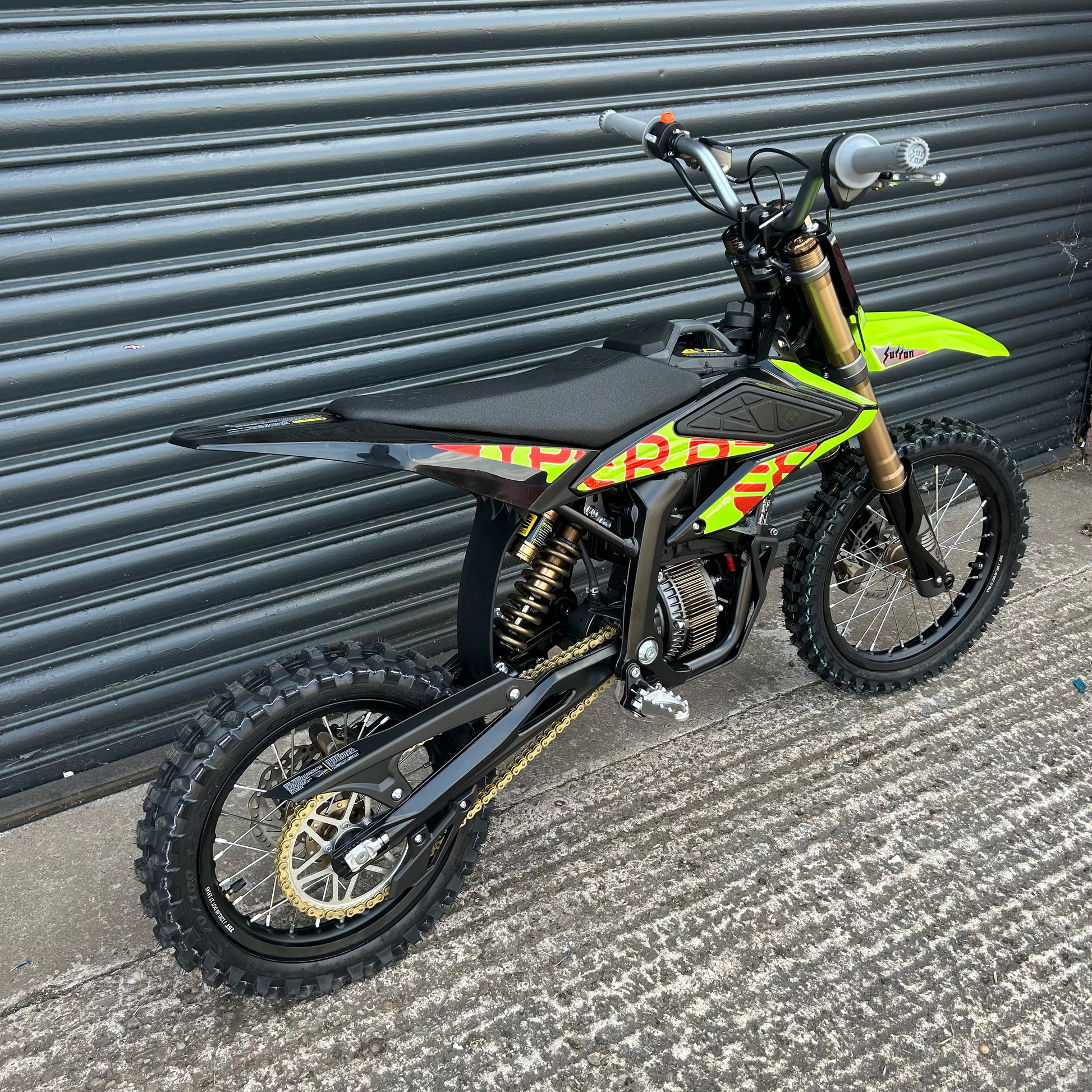 A black and lime green youth dirt bike with gold suspension forks and black knobby tires, parked against a dark gray corrugated metal garage door.