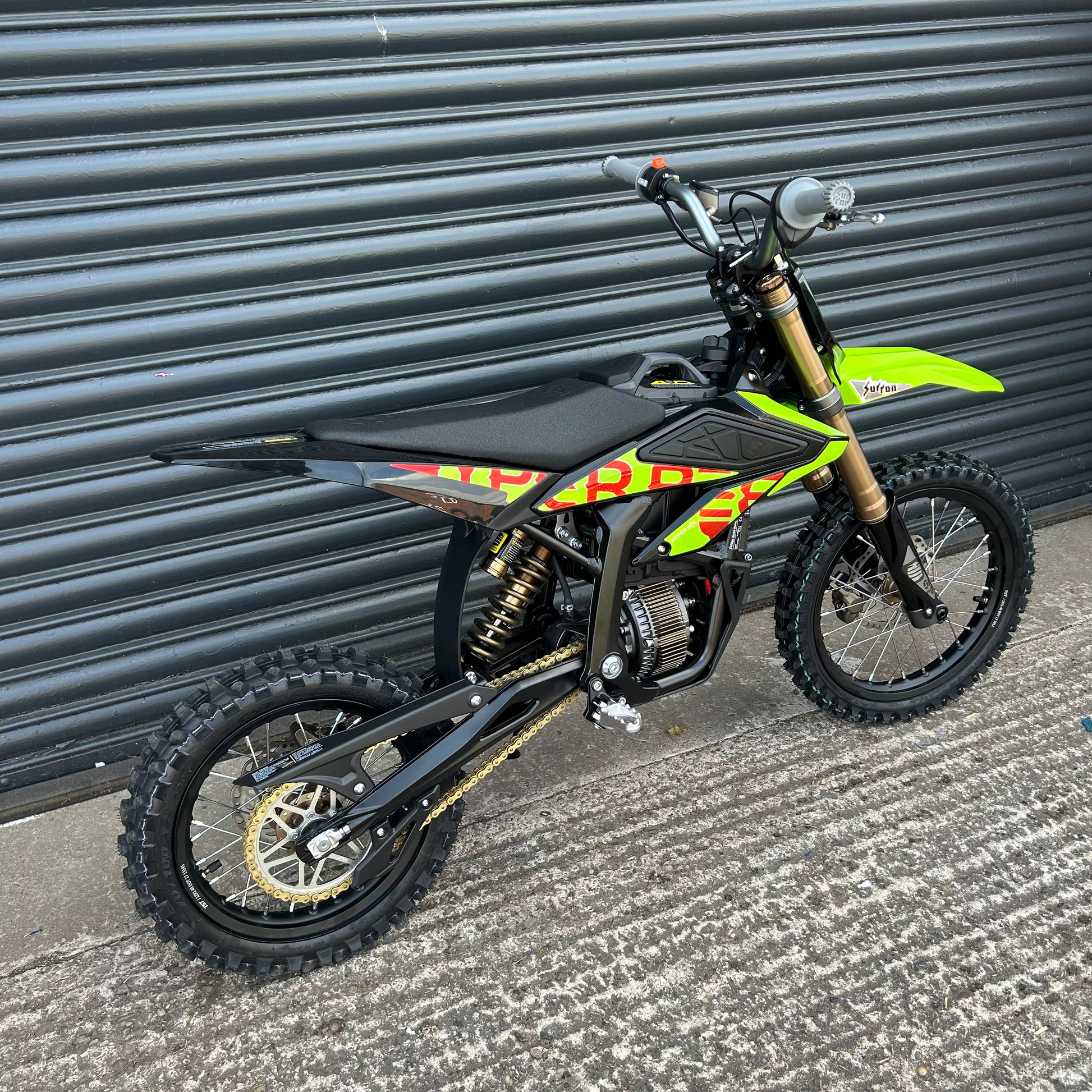 A small off-road dirt bike with a black seat and body, bright neon green fenders and side panel graphics, and gold front fork and rear suspension, photographed outdoors against a dark corrugated metal garage door.