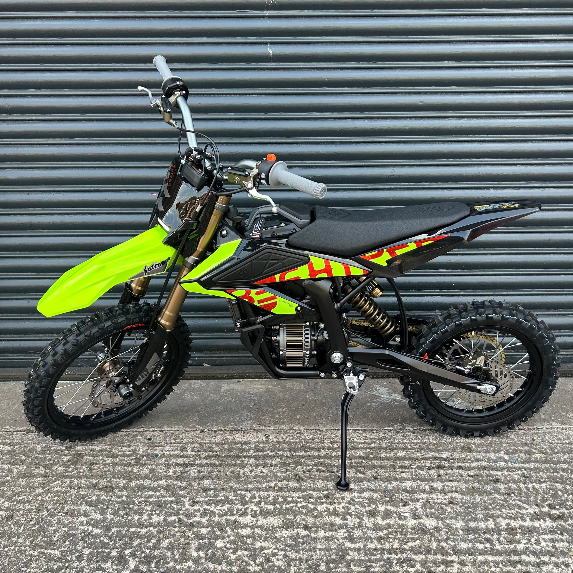 A small off-road dirt bike with a bright neon yellow-green front fender, black seat, and red and yellow graphic decals, parked on a concrete surface in front of a gray metal roll-up door.