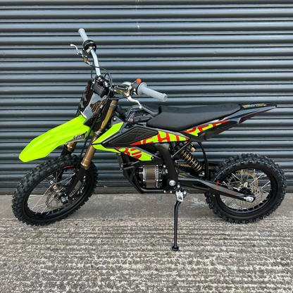 A small off-road dirt bike with a bright neon yellow-green front fender, black seat, and red and yellow graphic decals, parked on a concrete surface in front of a gray metal roll-up door.