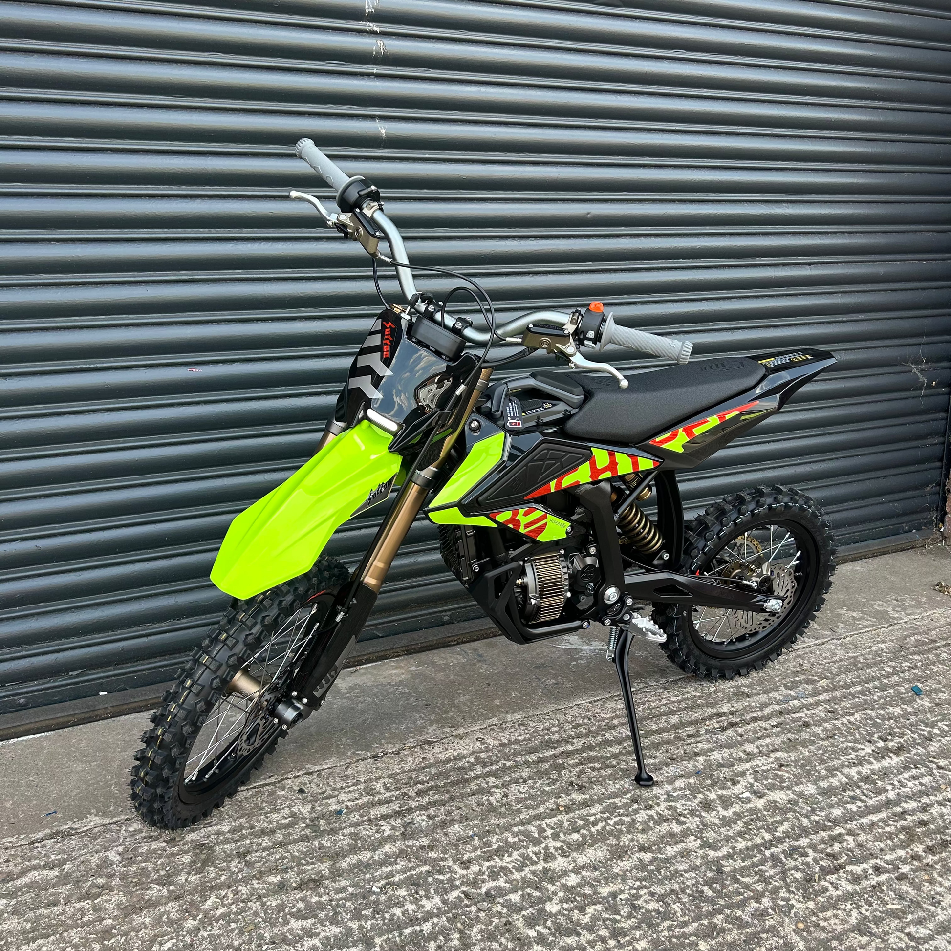 A small off-road dirt bike with a bright neon green front fender, black seat and body panels, and red/yellow graphic decals, photographed on a concrete surface in front of a dark gray metal garage door.