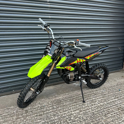 A small off-road dirt bike with a bright neon green front fender, black seat and body panels, and red/yellow graphic decals, photographed on a concrete surface in front of a dark gray metal garage door.