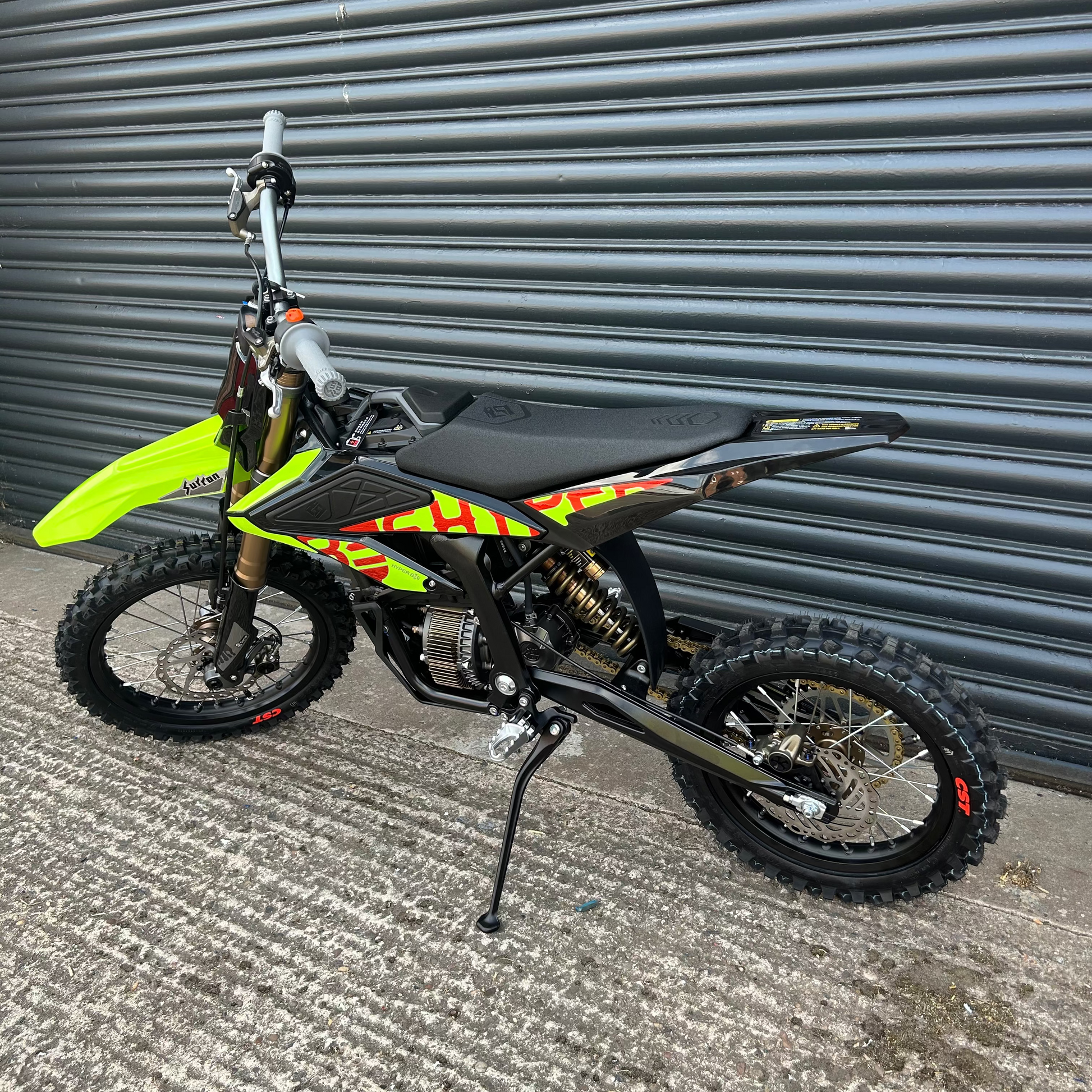 A youth dirt bike with a black seat and knobby tires, featuring a bright neon yellow-green front fender and orange/yellow graphic decals on the side, parked on concrete in front of a gray metal garage door.