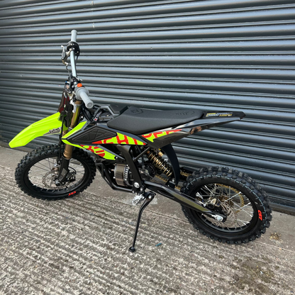 A youth dirt bike with a black seat and knobby tires, featuring a bright neon yellow-green front fender and orange/yellow graphic decals on the side, parked on concrete in front of a gray metal garage door.