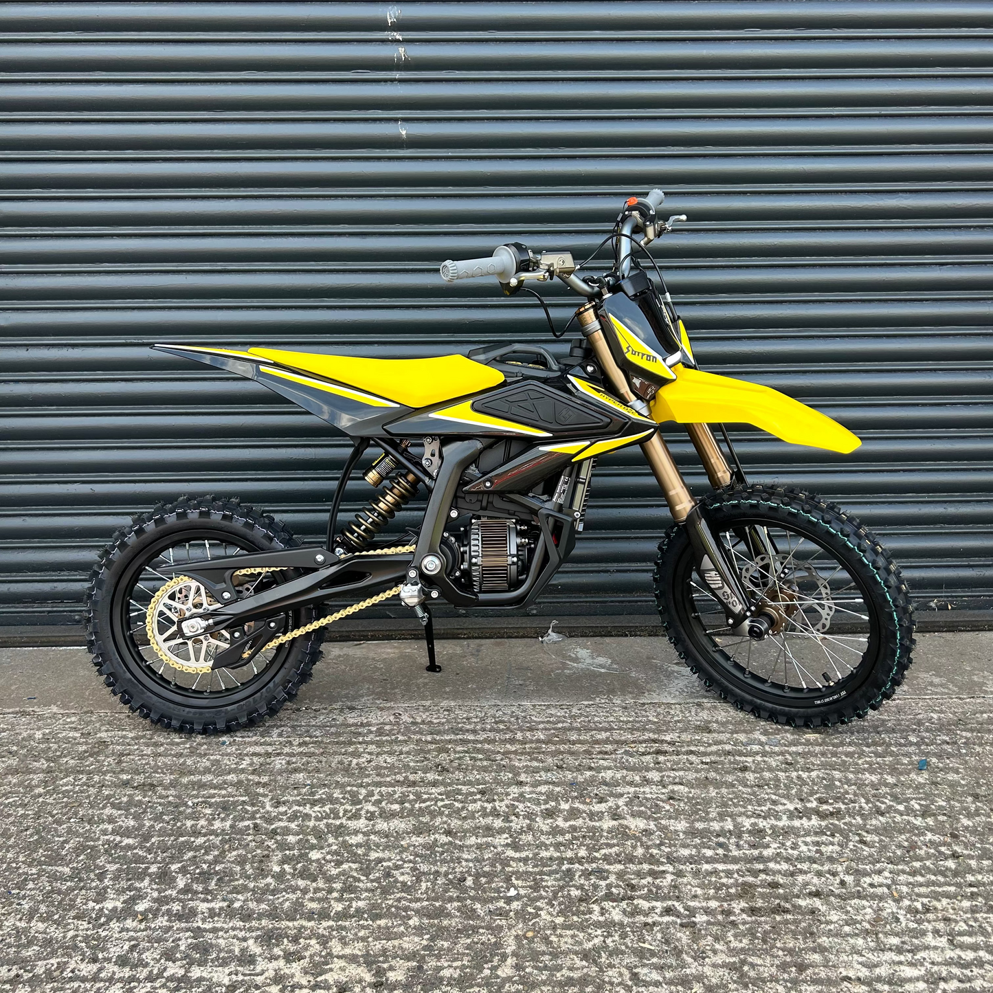 A yellow and black dirt bike with gold front fork details, parked on a concrete surface in front of a dark gray corrugated metal wall.