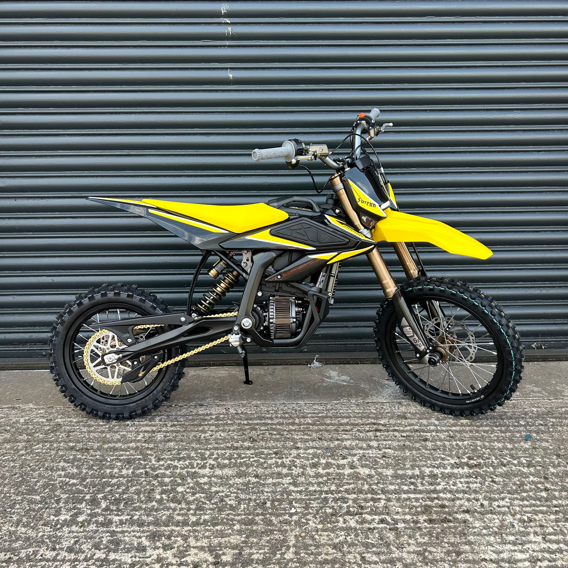 A yellow and black dirt bike with gold front fork details, parked on a concrete surface in front of a dark gray corrugated metal wall.