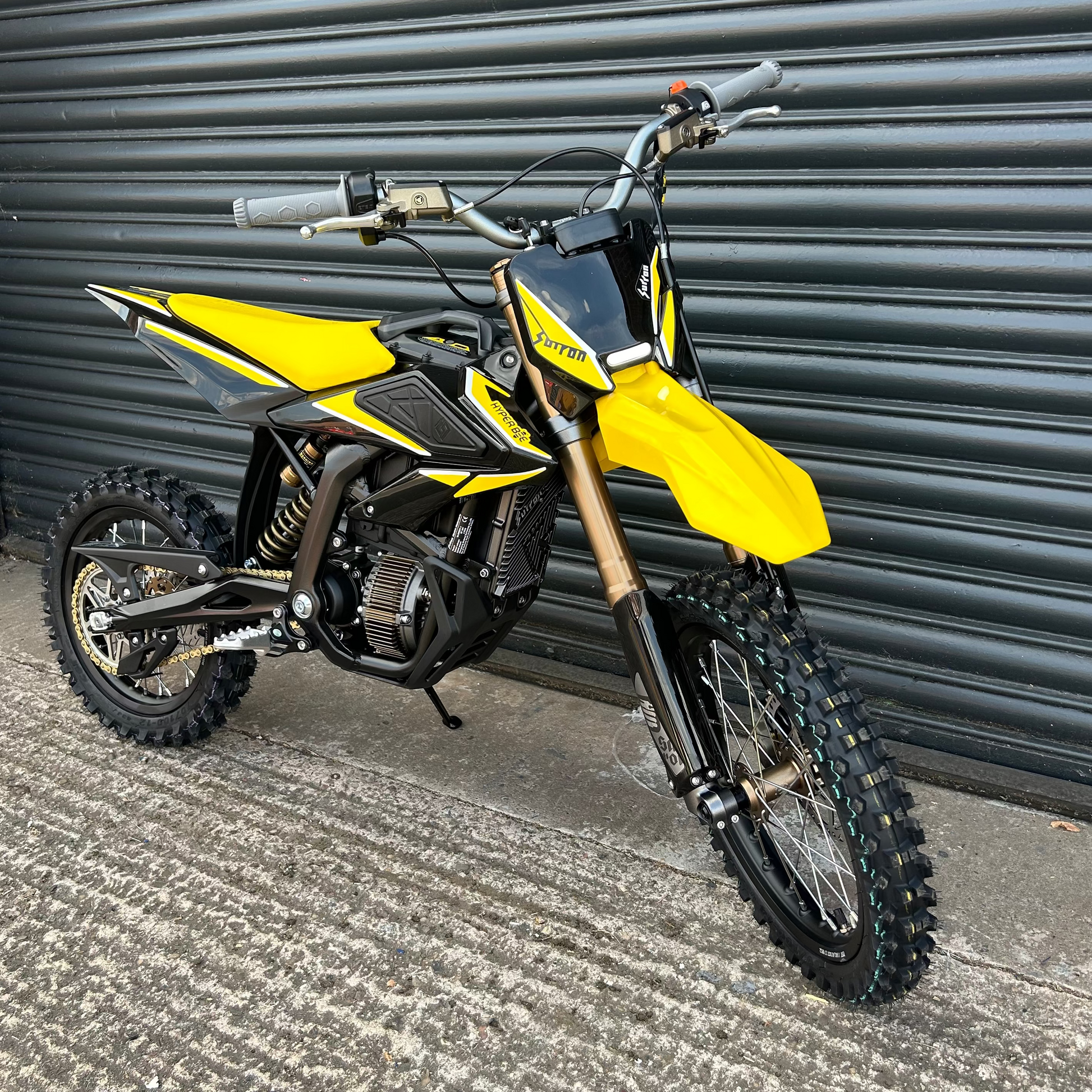 A yellow and black mini dirt bike with knobby tires, parked in front of a dark gray metal roller shutter door.