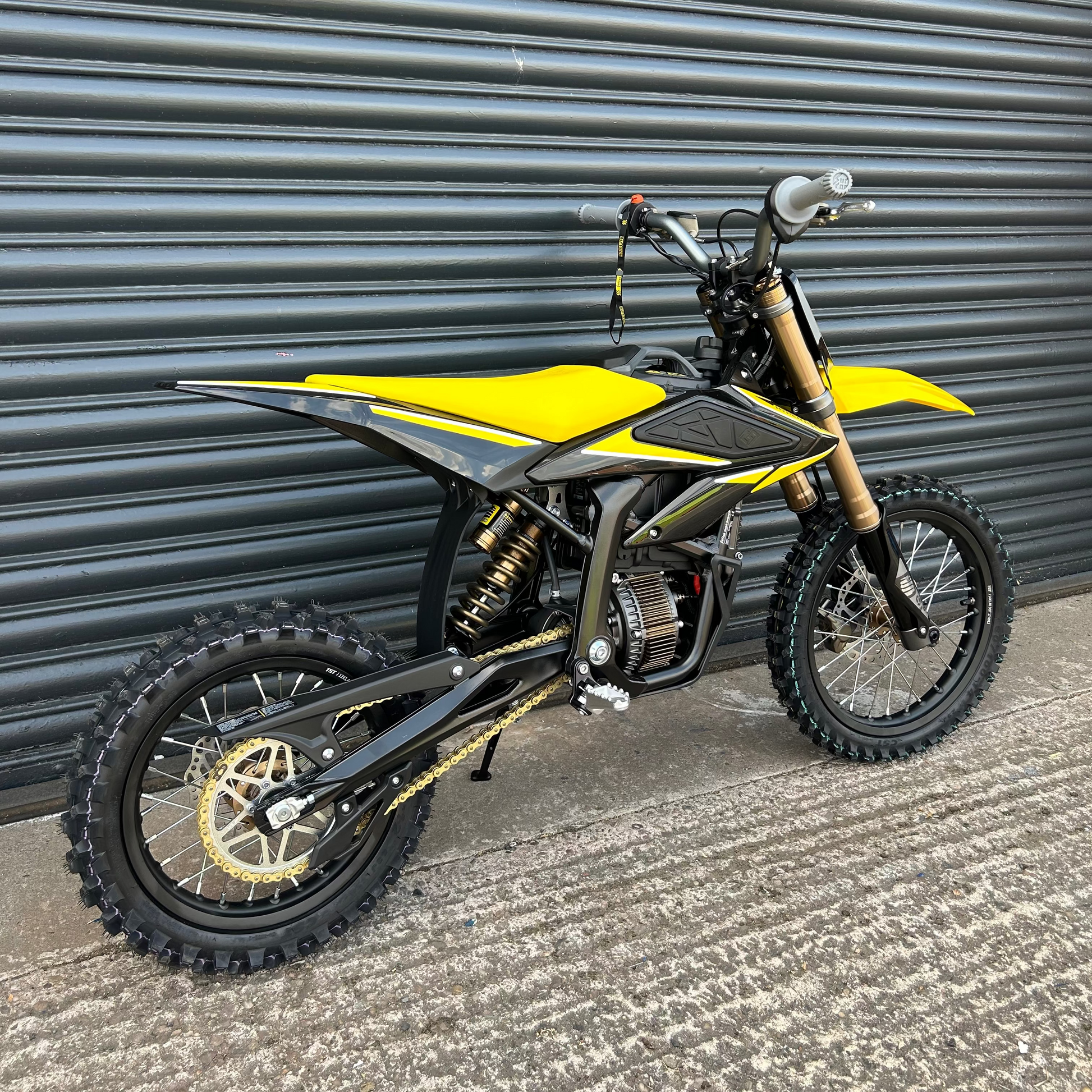 A yellow and black electric dirt bike with gold-colored front fork and rear wheel accents, parked against a dark gray corrugated metal garage door.