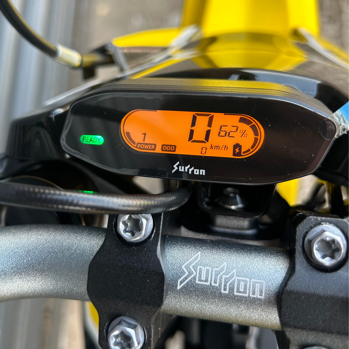 Close-up of a Surtron motorcycle dashboard display mounted on the handlebar, showing an orange digital screen and green battery indicator, with a yellow bike frame in the background.