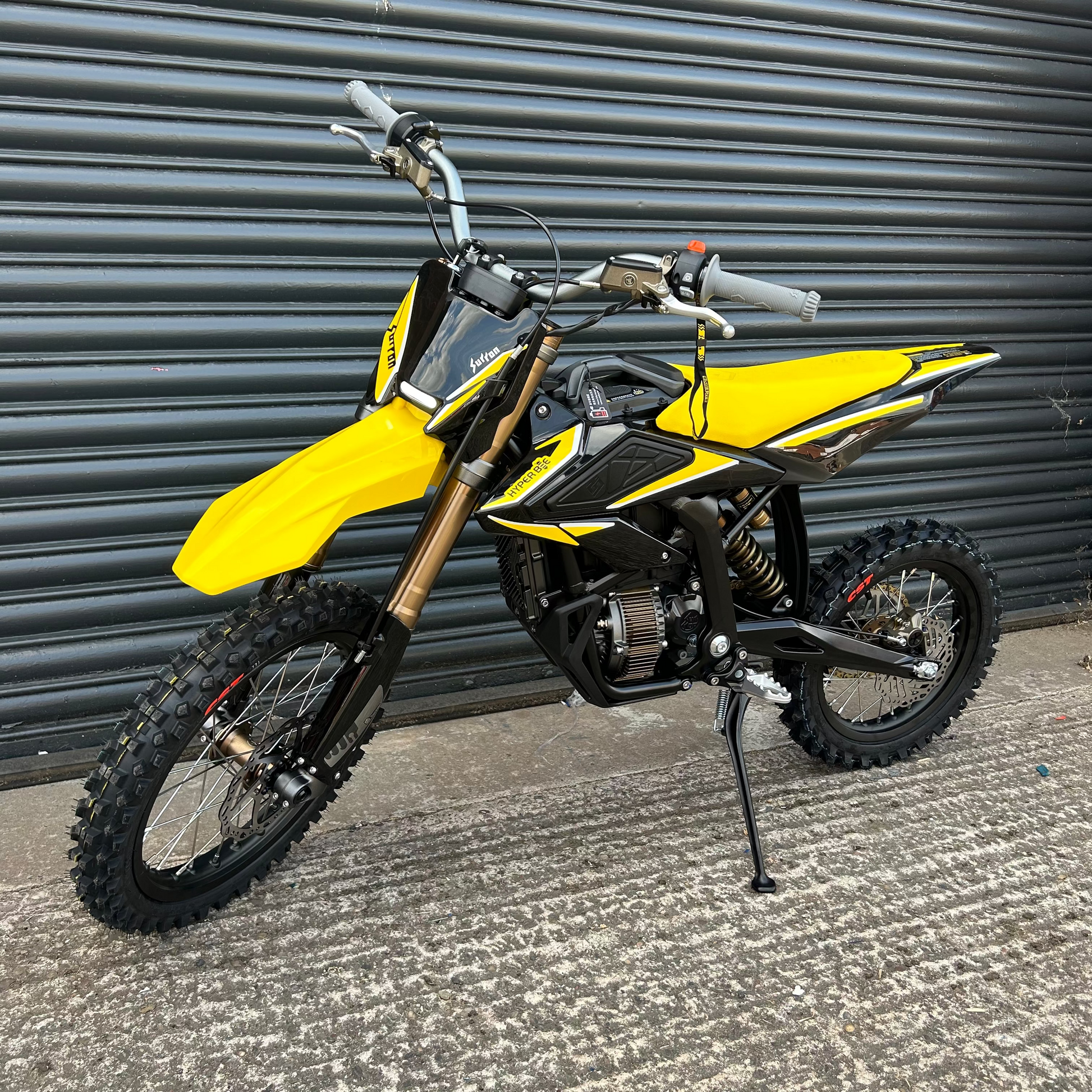 A yellow and black dirt bike with knobby off-road tires, standing on its kickstand in front of a gray metal roll-up door.