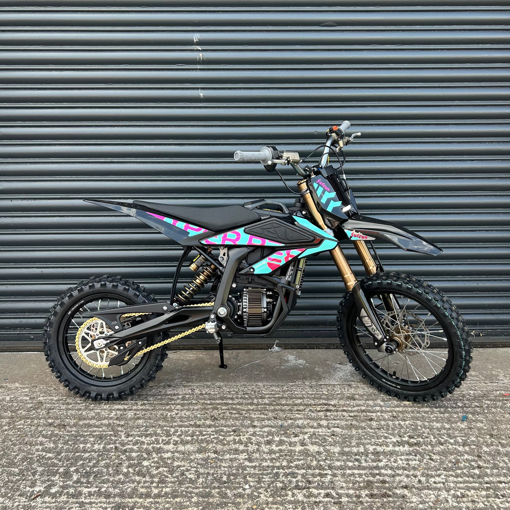 A black dirt bike with colorful pink, blue, and purple graphics, gold forks, and knobby tires, parked in front of a gray metal roll-up door.