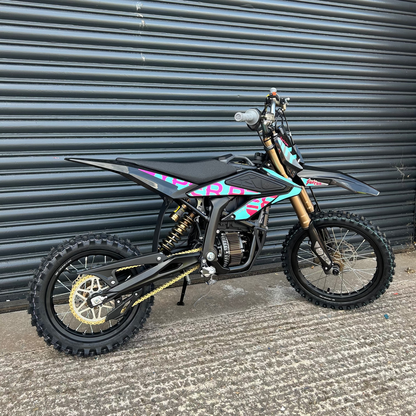 A black dirt bike with turquoise, pink, and purple graphic decals, gold forks, and black knobby tires, parked in front of a gray metal roller shutter door.