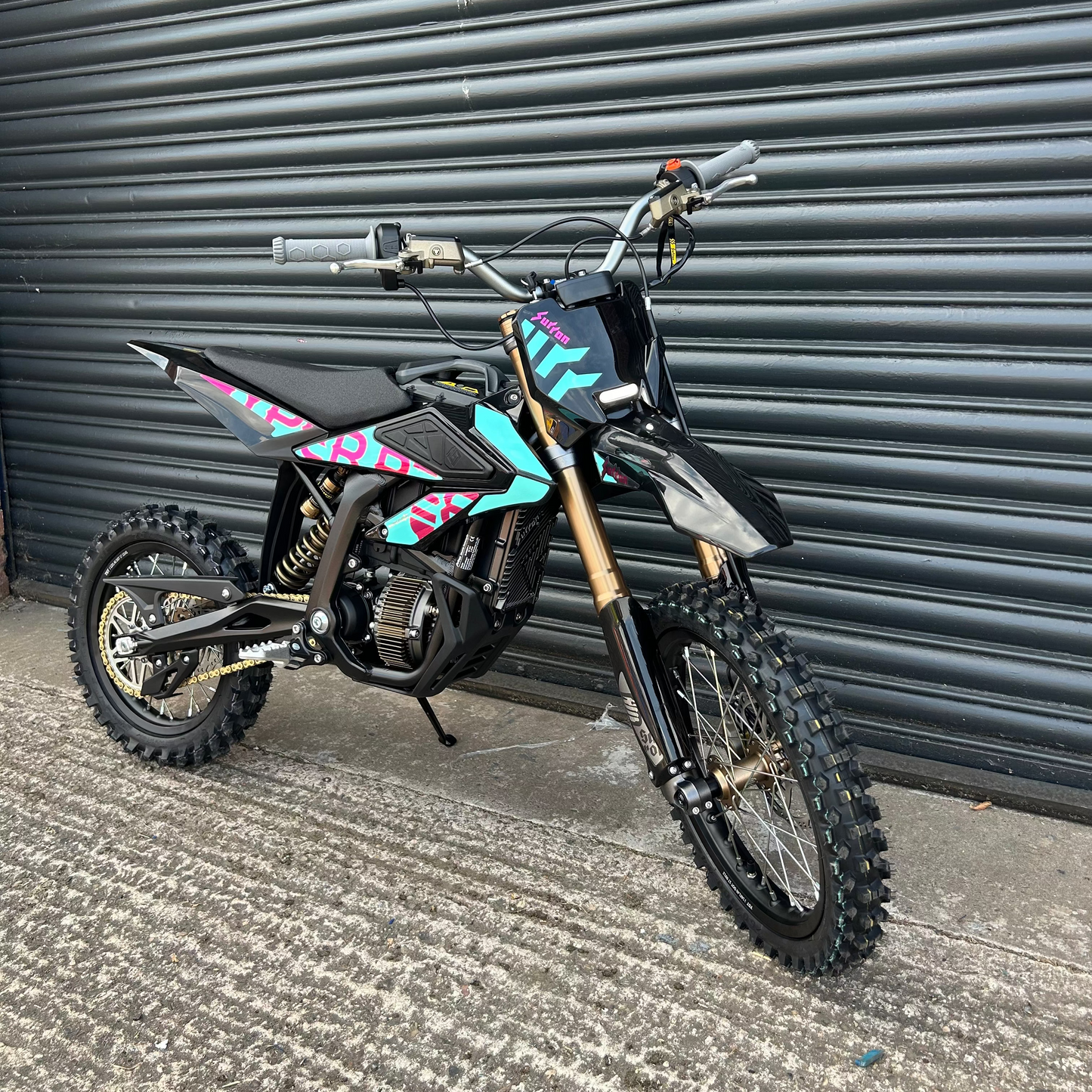 A small black dirt bike with colorful pink, blue, and teal graphics, gold front fork, and knobby tires, parked on concrete in front of a dark gray metal roll-up door.