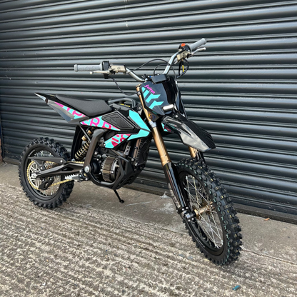 A small black dirt bike with colorful pink, blue, and teal graphics, gold front fork, and knobby tires, parked on concrete in front of a dark gray metal roll-up door.