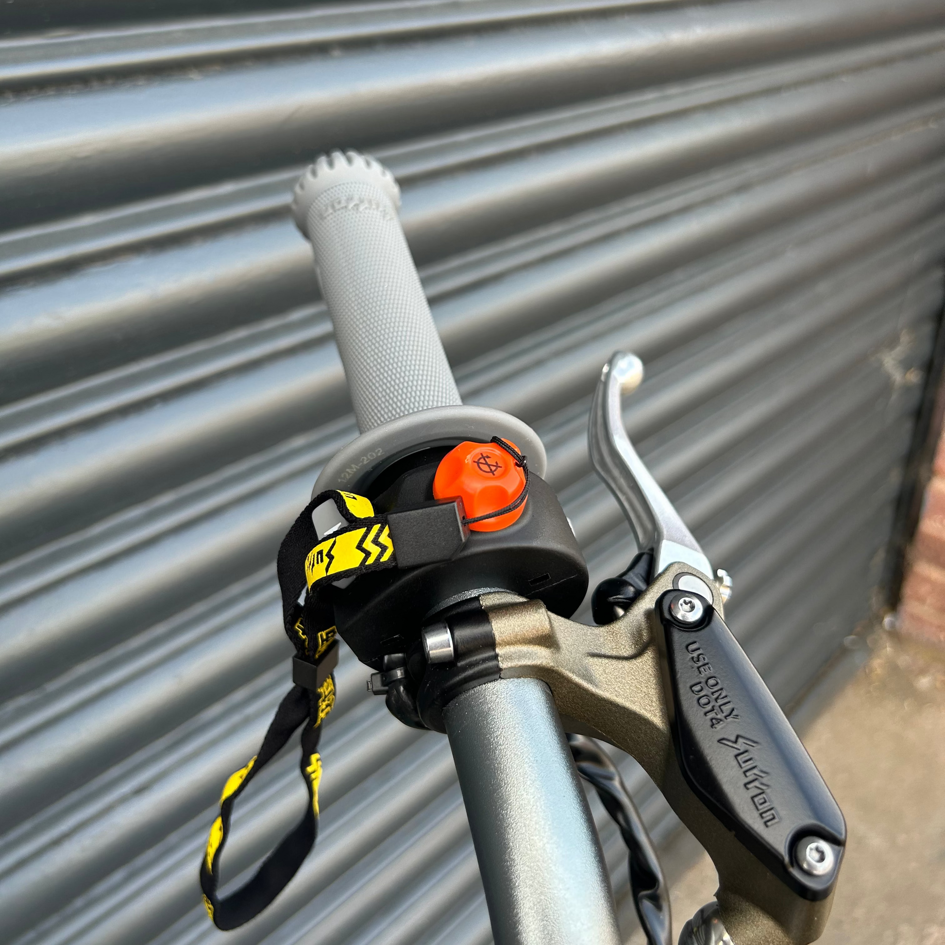 Close-up of a bicycle handlebar area with a black and yellow safety lanyard attached near the grip, alongside a brake lever, set against a gray metal shutter background.