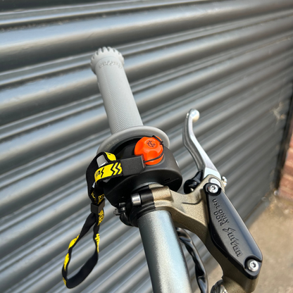 Close-up of a bicycle handlebar area with a black and yellow safety lanyard attached near the grip, alongside a brake lever, set against a gray metal shutter background.