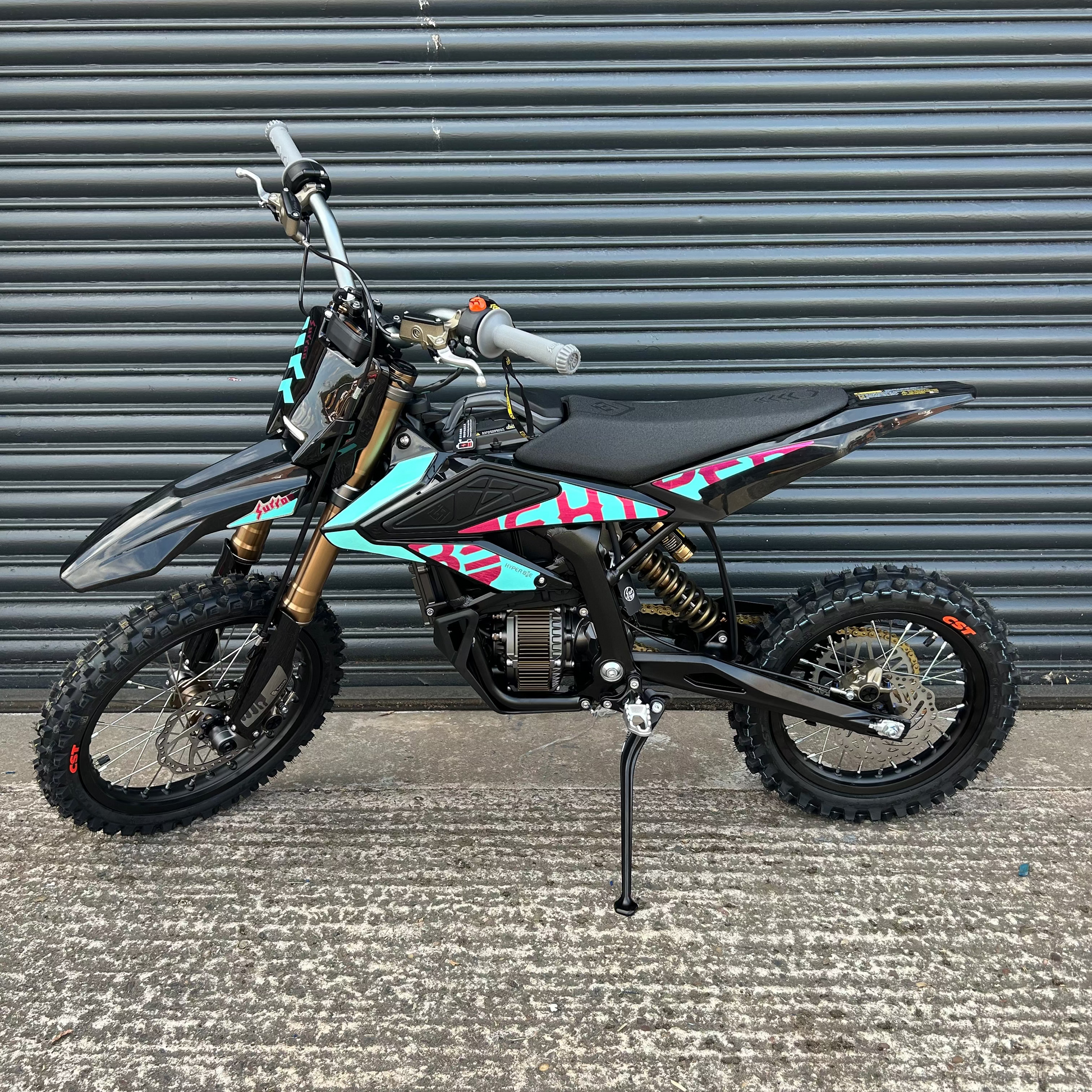 A small black dirt bike with turquoise and pink graphic decals, standing on its kickstand in front of a gray metal shutter door.
