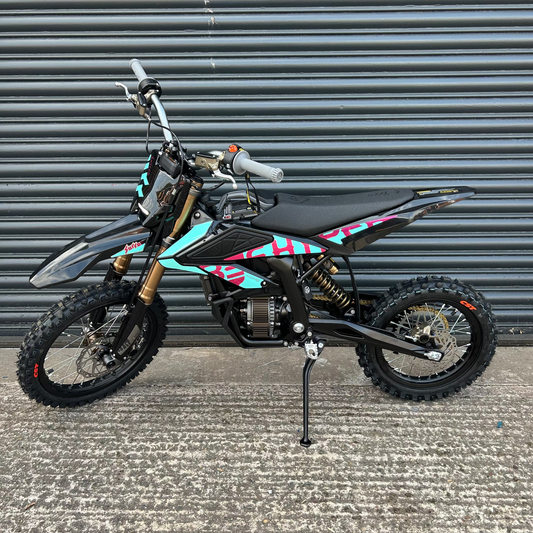 A small black dirt bike with turquoise and pink graphic decals, standing on its kickstand in front of a gray metal shutter door.