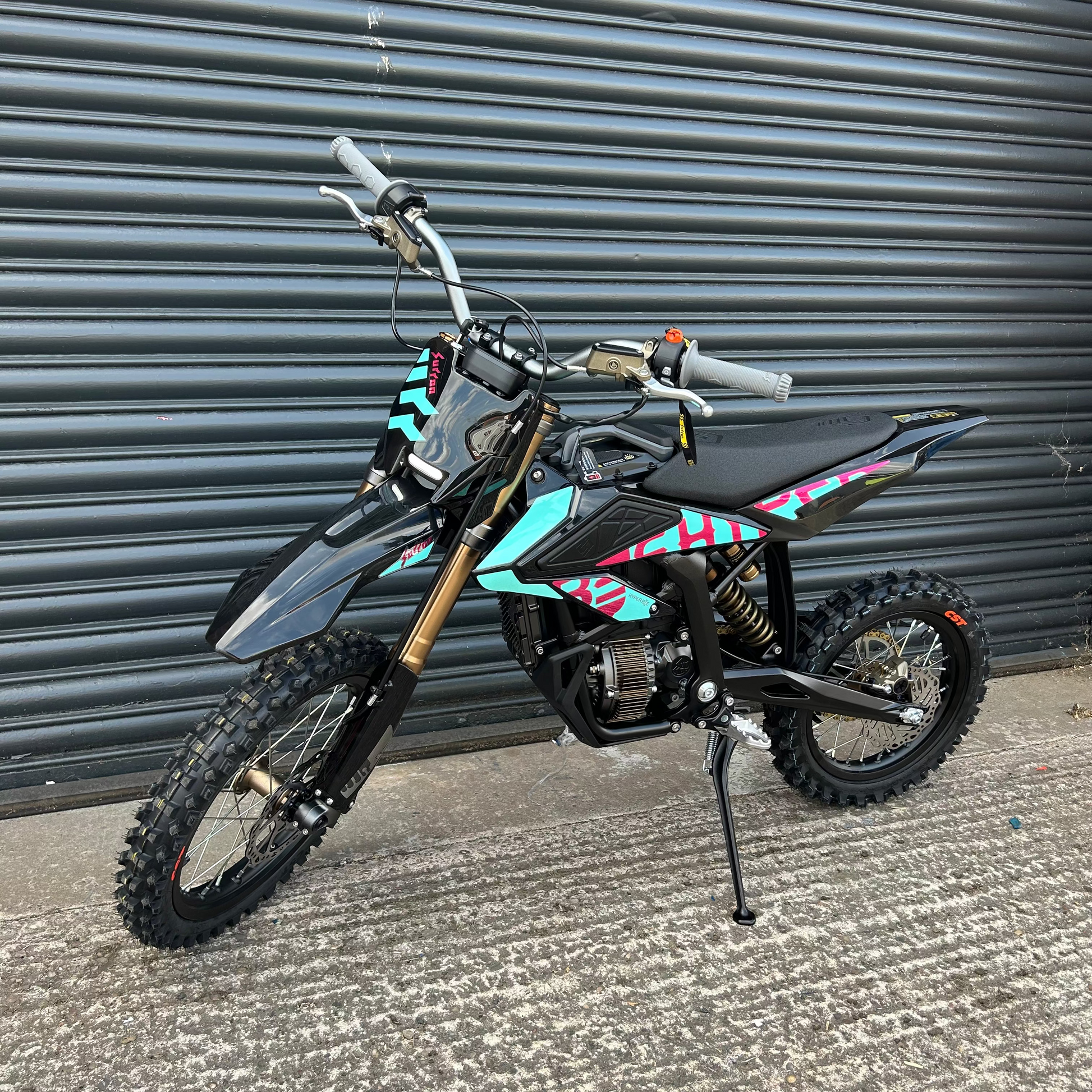 A small off-road dirt bike with black and gray bodywork, gold front fork, and turquoise and pink graphic decals, parked on concrete in front of a gray corrugated metal shutter.