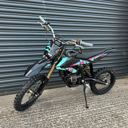 A small off-road dirt bike with black and gray bodywork, gold front fork, and turquoise and pink graphic decals, parked on concrete in front of a gray corrugated metal shutter.