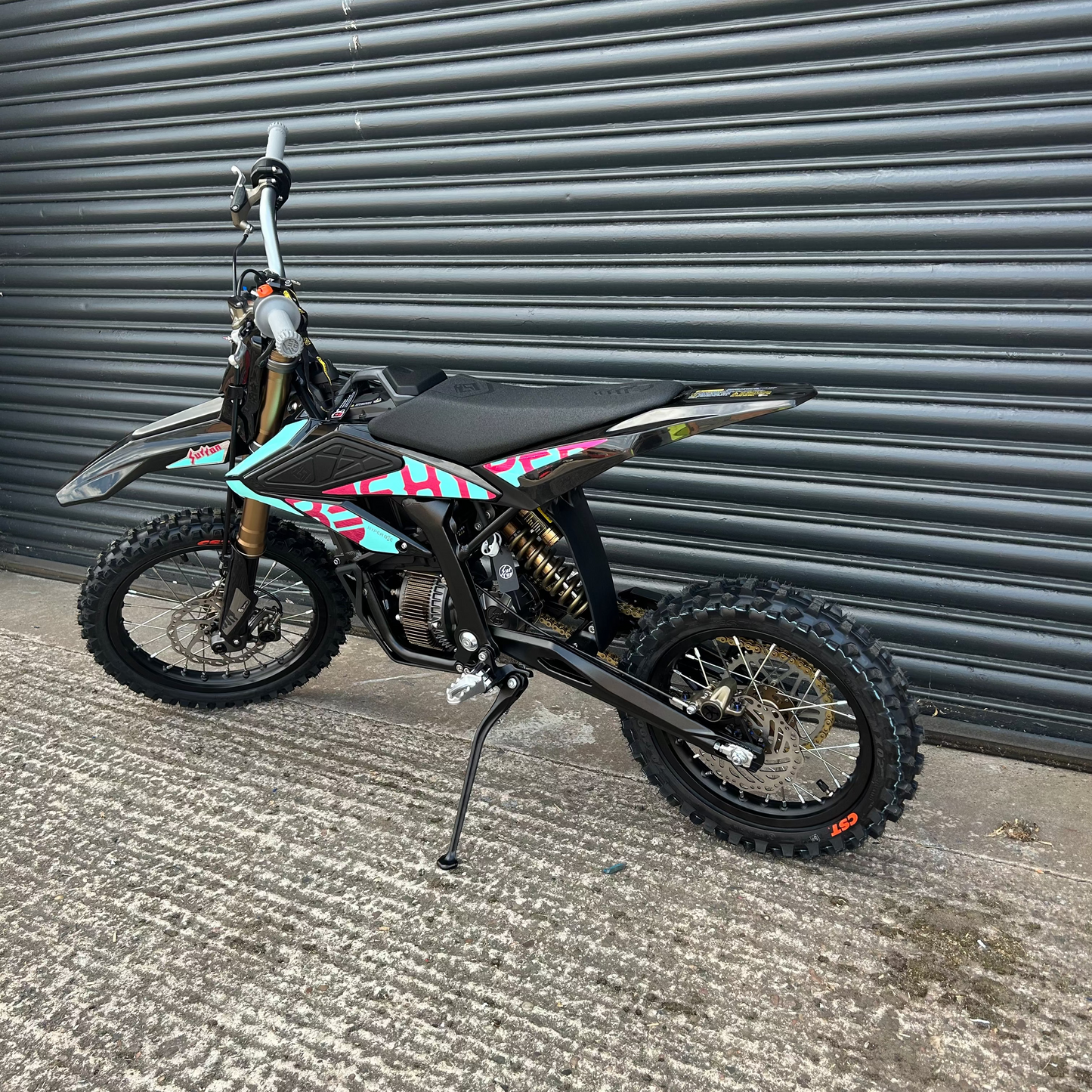 A black dirt bike with colorful geometric decals in pink, teal, and white, parked on a concrete surface in front of a gray metal roller shutter door.
