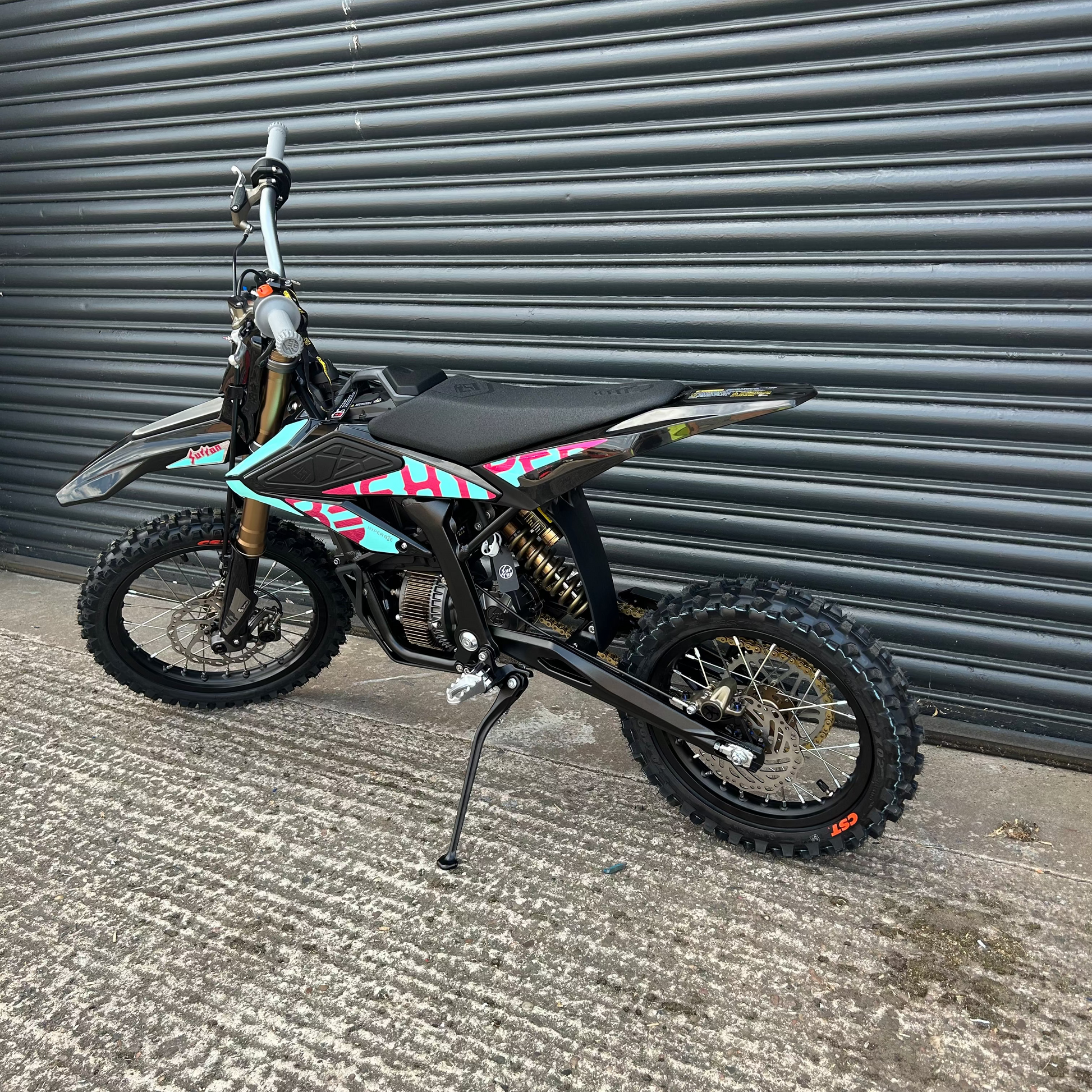 A black dirt bike with colorful geometric decals in pink, teal, and white, parked on a concrete surface in front of a gray metal roller shutter door.