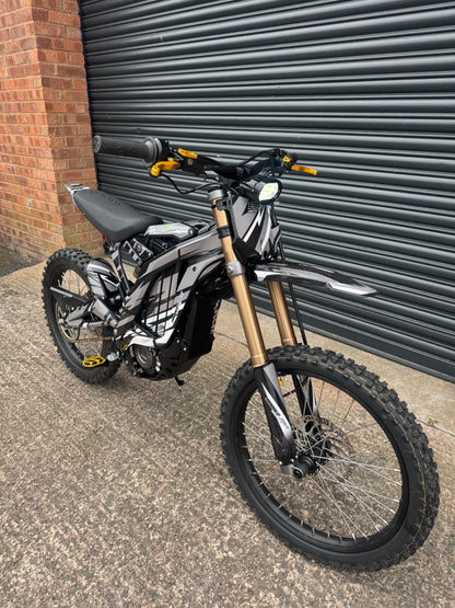 A black and silver electric dirt bike with gold front forks, parked on concrete in front of a dark gray metal roll-up door and a brick wall.