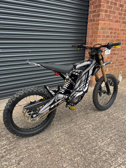 A black and silver electric dirt bike with a gold front fork, parked on concrete next to a brick wall and a dark metal garage door.