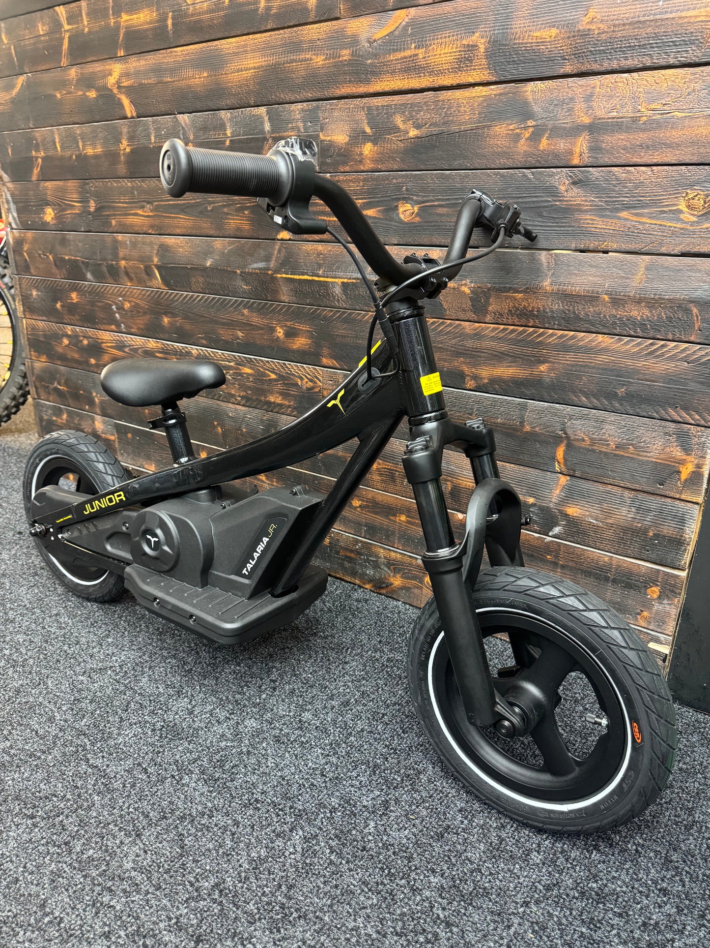 A black electric kids' balance bike with a seat and front suspension fork, photographed indoors against a dark wood plank wall on a gray textured floor.