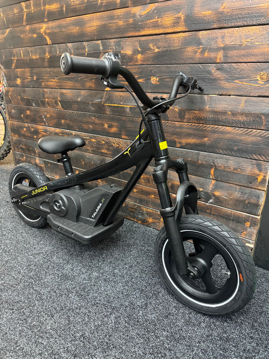 A black electric kids' balance bike with a seat and front suspension fork, photographed indoors against a dark wood plank wall on a gray textured floor.