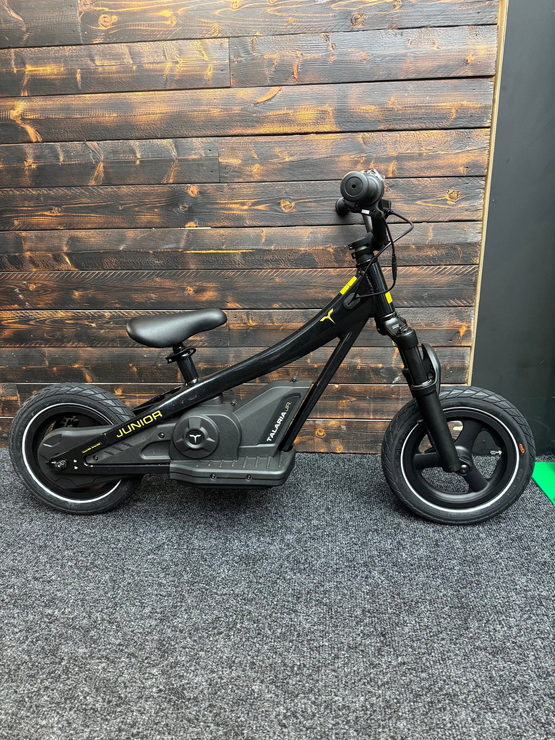 A black electric kids' balance bike with a step-through frame, front suspension fork, and rear wheel motor hub, shown against a dark wood plank wall.