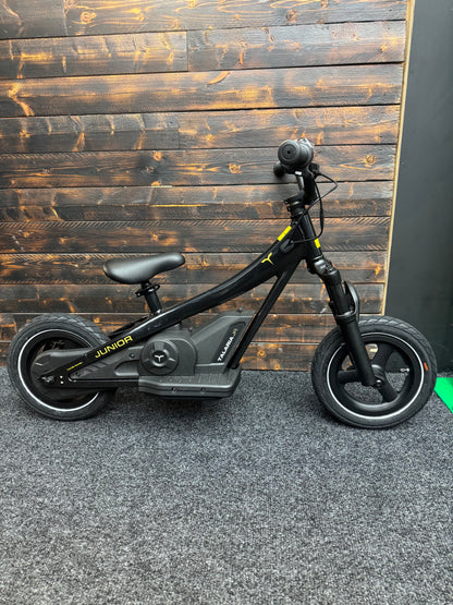 A black electric kids' balance bike with a step-through frame, front suspension fork, and rear wheel motor hub, shown against a dark wood plank wall.