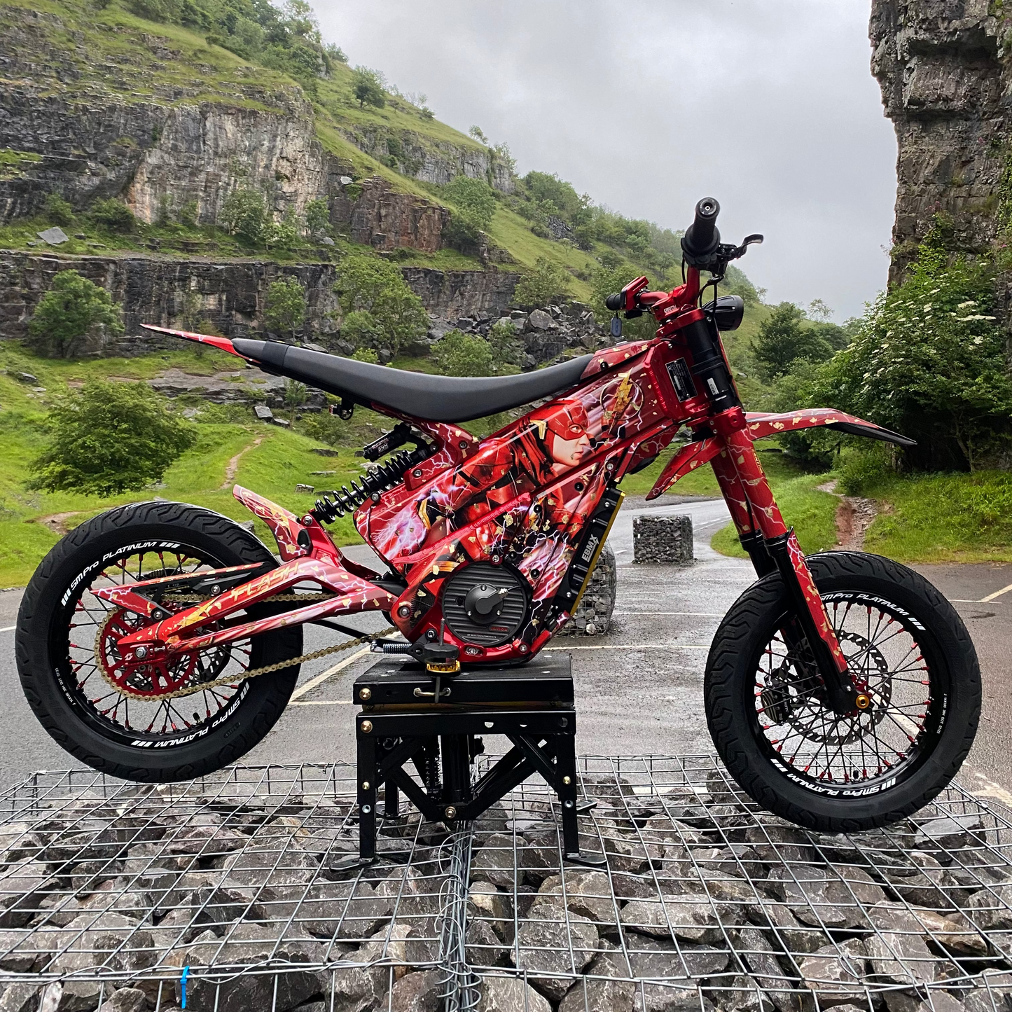 A red and black custom electric dirt bike with a graphic wrap, displayed on a black stand in an outdoor rocky mountain setting.