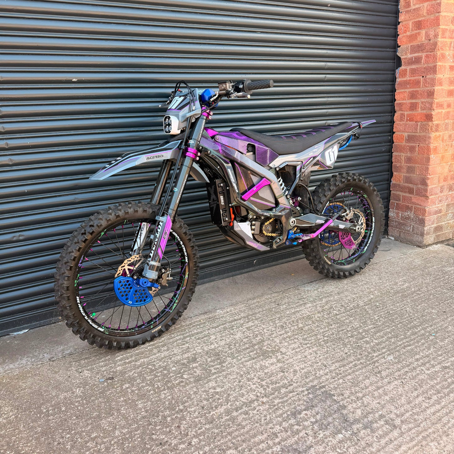 A custom electric dirt bike with a black and purple body, custom titanium exhaust, and blue brake disc, parked against a dark gray metal garage door.