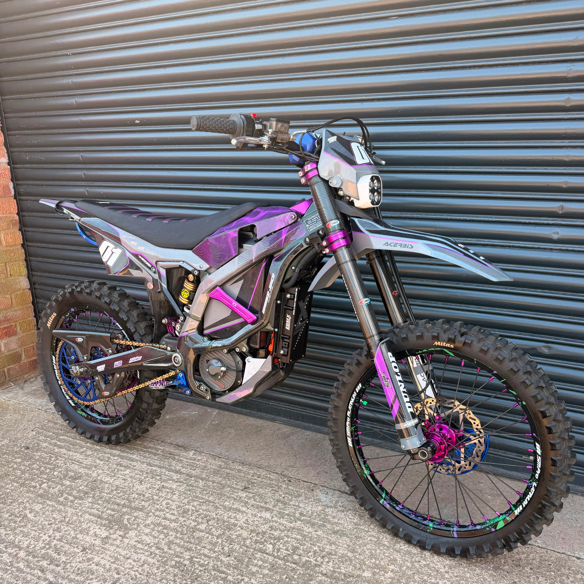 A custom electric dirt bike with a black, purple, and silver frame, parked against a gray metal garage door.