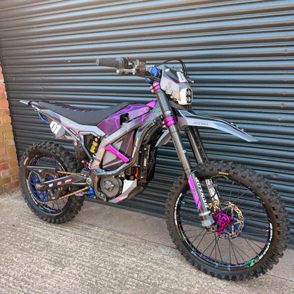 A custom electric dirt bike with a black, purple, and silver frame, parked against a gray metal garage door.