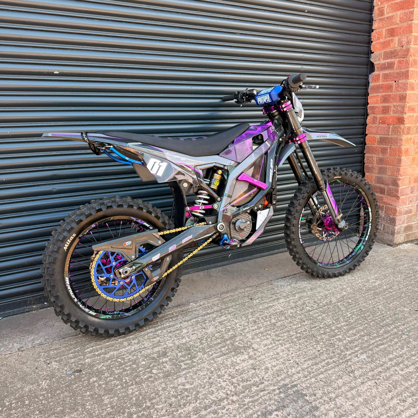 A custom electric dirt bike with a metallic purple and silver body, black seat, and colorful blue, gold, and purple accents, parked against a black metal garage door next to a brick wall.