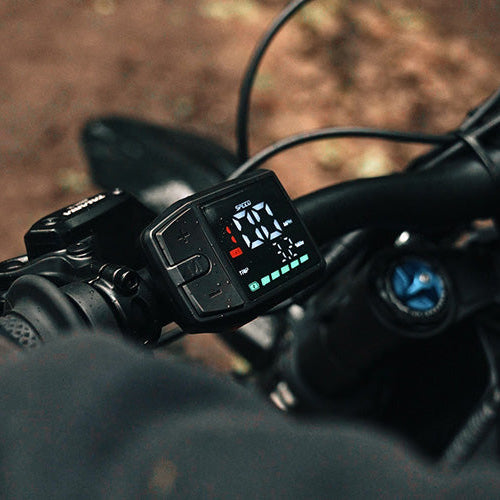 Close-up of a black electronic bike display mounted on a handlebar, showing a digital speed readout, with part of a rider's gloved hand visible on the left and a blurred outdoor ground background.
