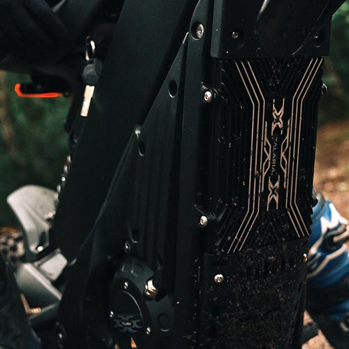 Close-up of a black metal bicycle frame section with decorative gold line patterns, photographed outdoors on a dirt trail with a rider's leg visible in blue and white gear.